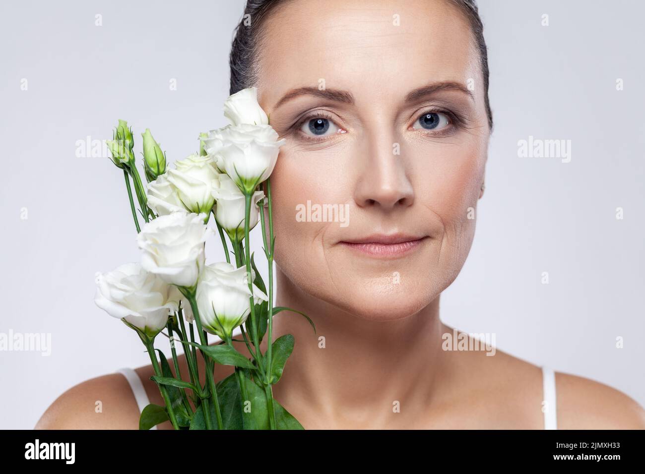Retrato de primer plano de hermosa mujer de mediana edad con una piel perfecta sosteniendo flores, mirando la cámara con expresión tranquila. Estudio de interior grabado aislado sobre fondo gris. Foto de stock