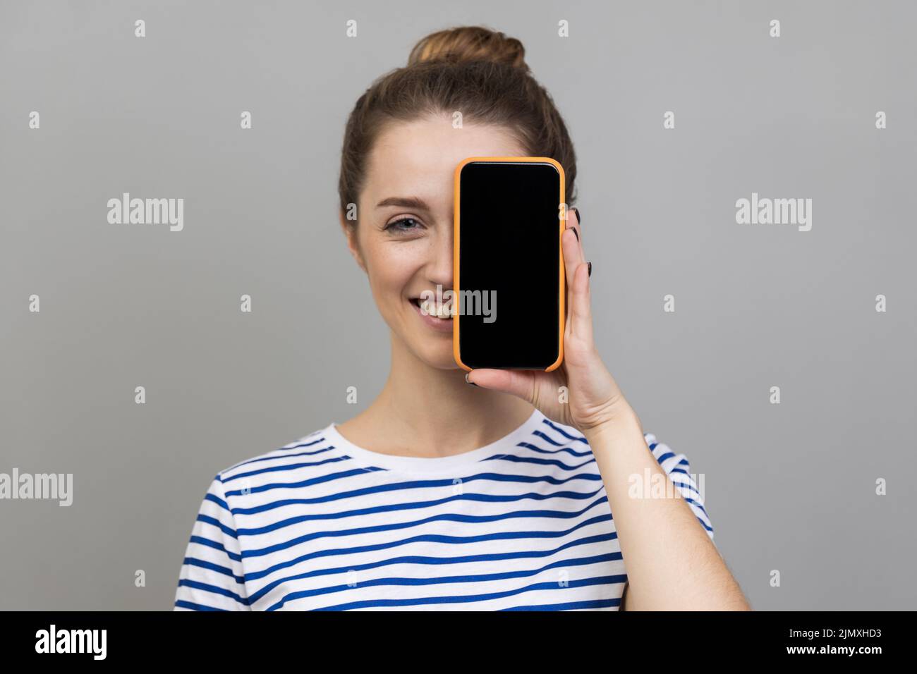 Retrato de atractiva mujer satisfecha con una camiseta a rayas que cubre el ojo con teléfono móvil con pantalla vacía para publicidad. Estudio de interior grabado aislado sobre fondo gris. Foto de stock