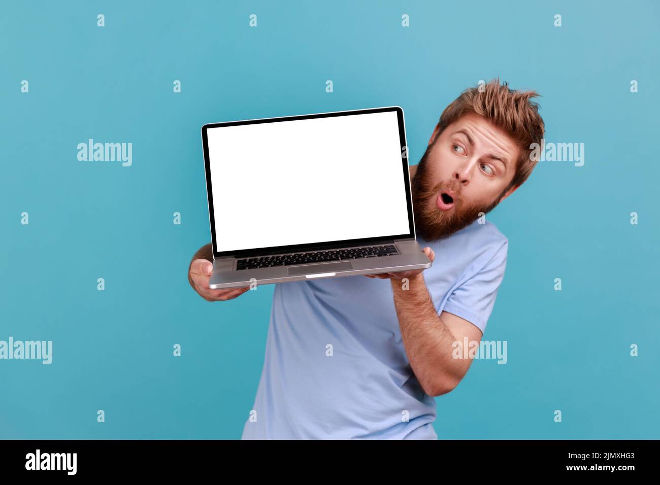 Retrato de un hombre barbudo sosteniendo un portátil con pantalla blanca en blanco y mirando la pantalla desde el lado con una expresión facial impactante, diciendo wow. Estudio de interior aislado sobre fondo azul. Foto de stock