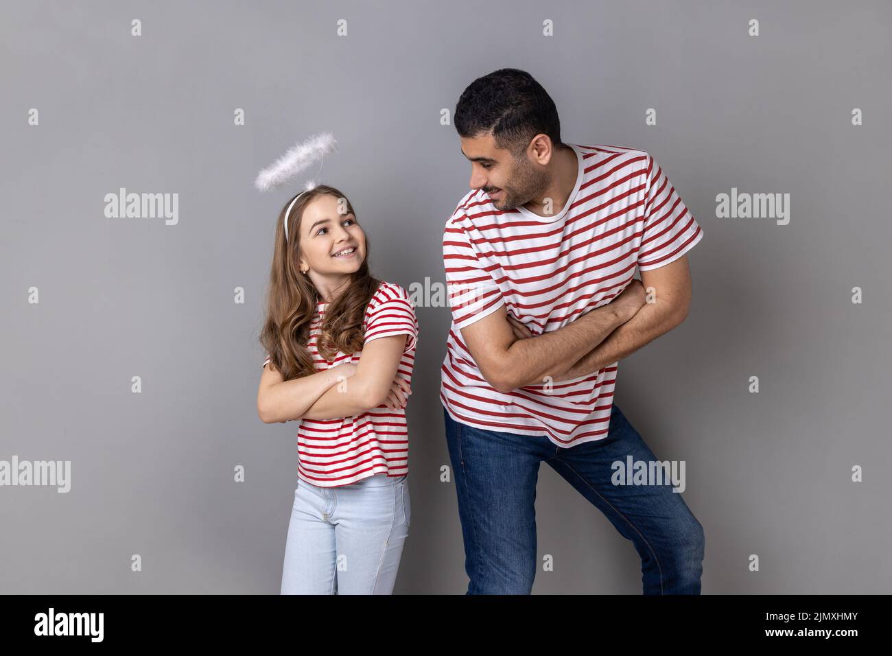 Retrato de un padre y una hija alegres y sonrientes en camisetas a rayas de pie con las manos dobladas y mirándose, la familia expresando la felicidad. Estudio de interior grabado aislado sobre fondo gris. Foto de stock