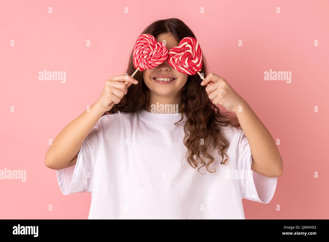 Retrato de niña con una camiseta blanca que cubre los ojos con caramelos en forma de corazón y sonriendo alegremente, posando con los labios de la puta. Estudio de interior grabado aislado sobre fondo rosa. Foto de stock