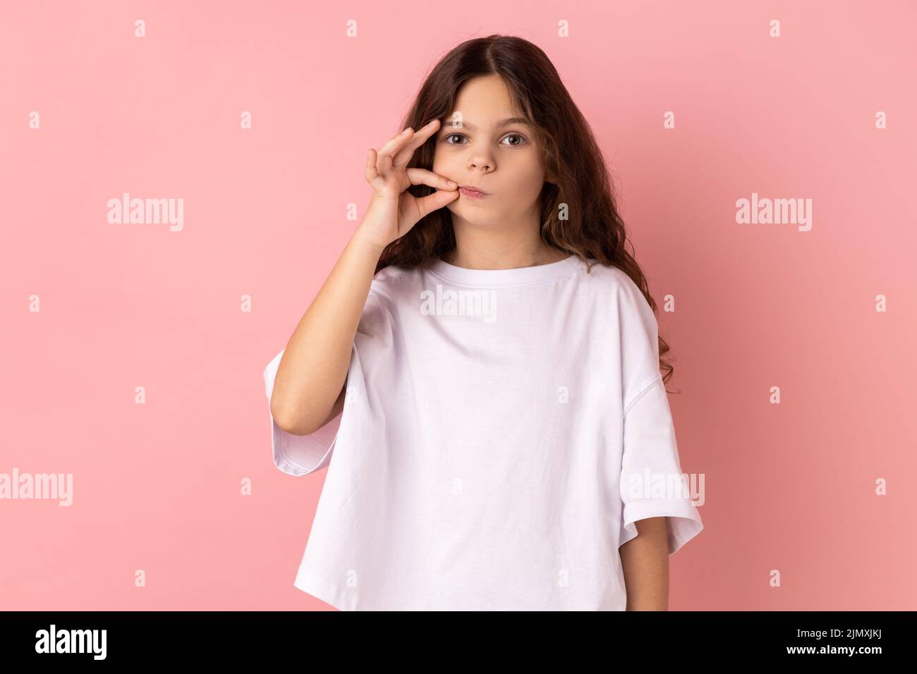 Retrato de una encantadora niña con una camiseta blanca con aspecto misterioso haciendo un gesto de cremallera para cerrar la boca, manteniendo el secreto, rematando los labios. Estudio de interior grabado aislado sobre fondo rosa. Foto de stock
