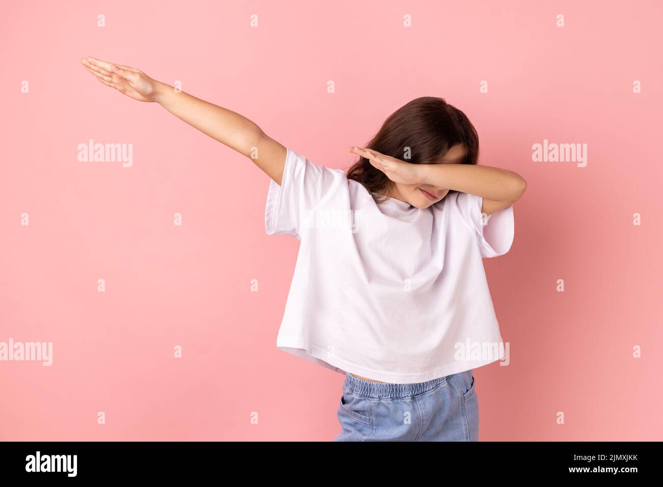 Retrato de niña con camiseta blanca que muestra la pose de baile de dab, el famoso meme de triunfo de Internet, realizando tendencias diabólicas con gesto de la mano. Estudio de interior grabado aislado sobre fondo rosa. Foto de stock