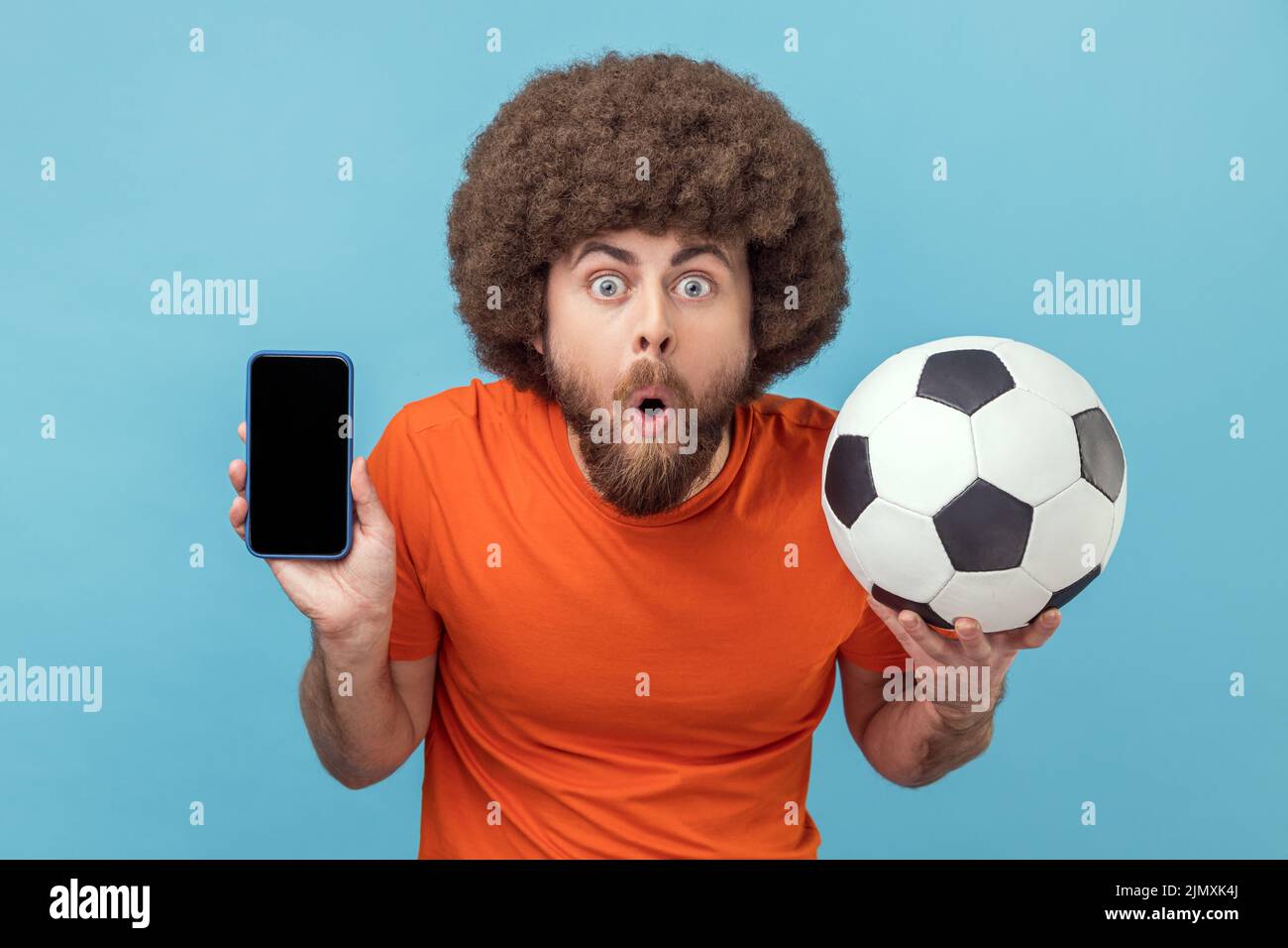 Retrato de hombre conmocionado con peinado afro usando camiseta de pie con balón de fútbol y mostrando teléfono celular con pantalla vacía para el anuncio. Estudio de interior aislado sobre fondo azul. Foto de stock
