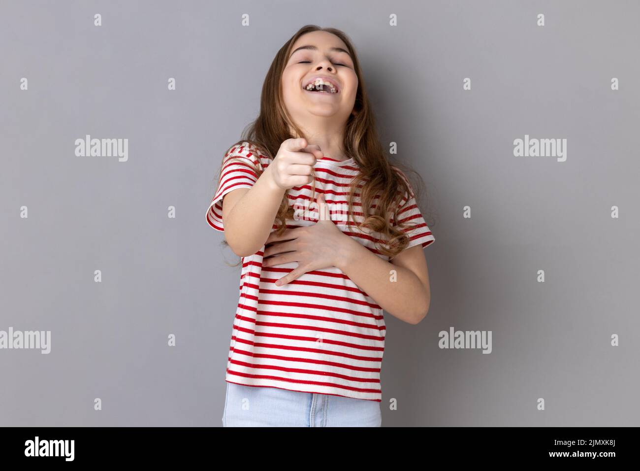 Retrato de niña que llevaba una camiseta a rayas riendo, sosteniendo el estómago y señalando a la cámara, burlándose de ti, no puede detener la risa histérica. Estudio de interior grabado aislado sobre fondo gris. Foto de stock