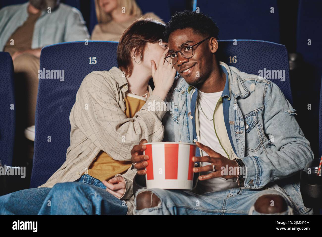 Joven mujer caucásica susurrando algo en el oído de su novio negro mientras ve una película en el cine Foto de stock