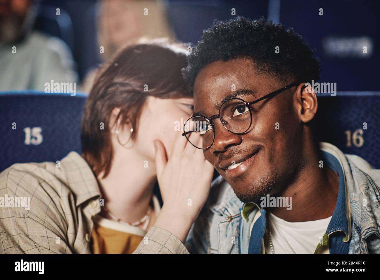 Primer plano de una joven mujer caucásica susurrando algo a su amiga negra mientras ve una película en el cine Foto de stock