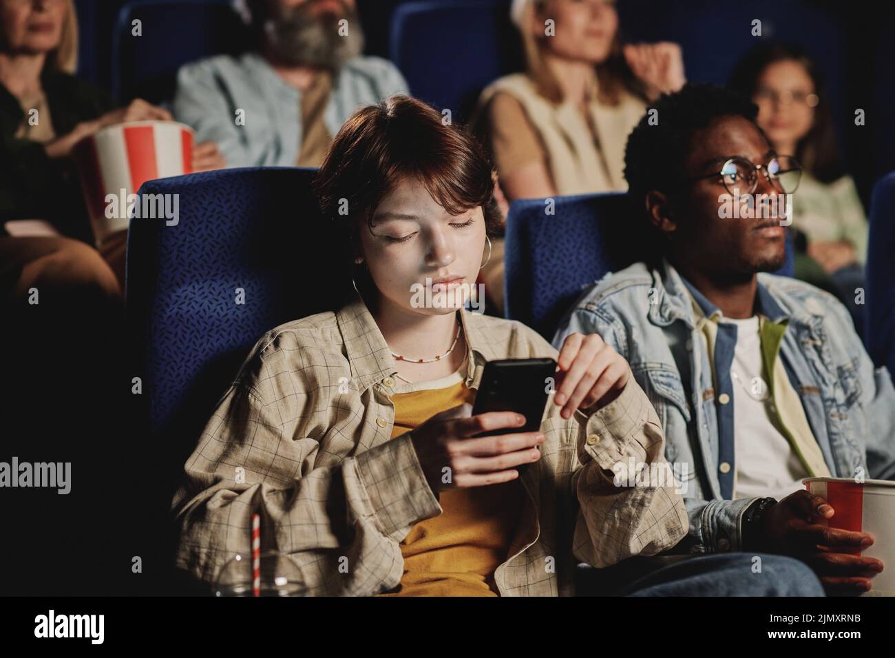 Joven gen Z mujer caucásica aburrirse con películas de desplazamiento de noticias en la red social en smartphone en el cine Foto de stock