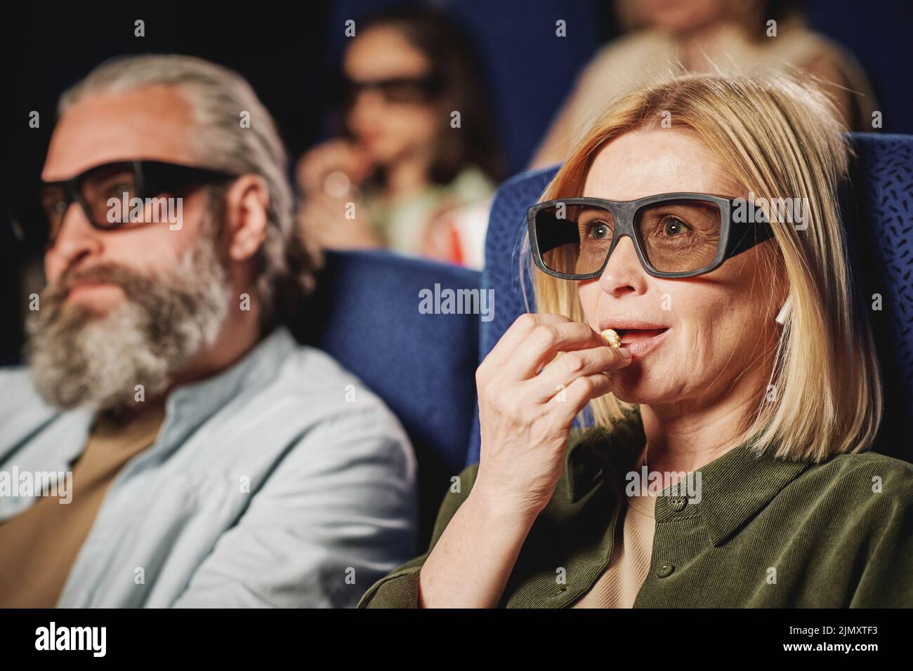 Foto de enfoque selectivo de mujer caucásica madura con pelo rubio, con gafas 3D viendo una película y comiendo palomitas en el cine Foto de stock