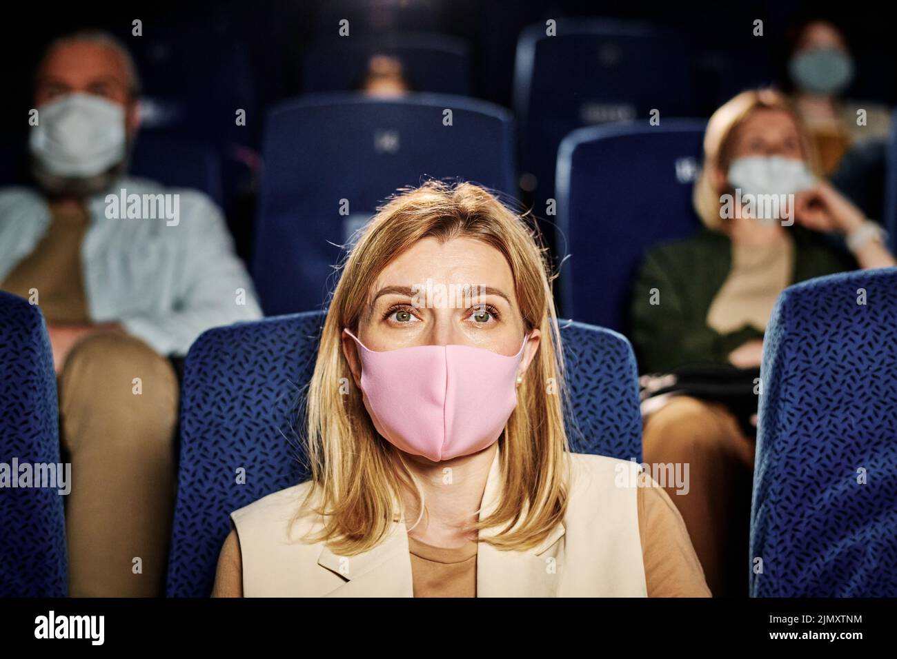 Retrato de mujer caucásica con máscara protectora rosa viendo una película en el cine, concepto de pandemia Foto de stock