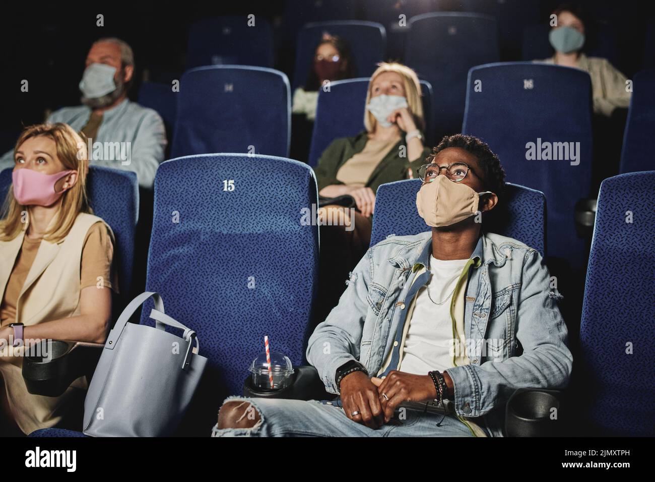 Grupo étnicamente diverso de personas que usan máscaras protectoras en las caras viendo películas en el cine, concepto de cuarentena Foto de stock