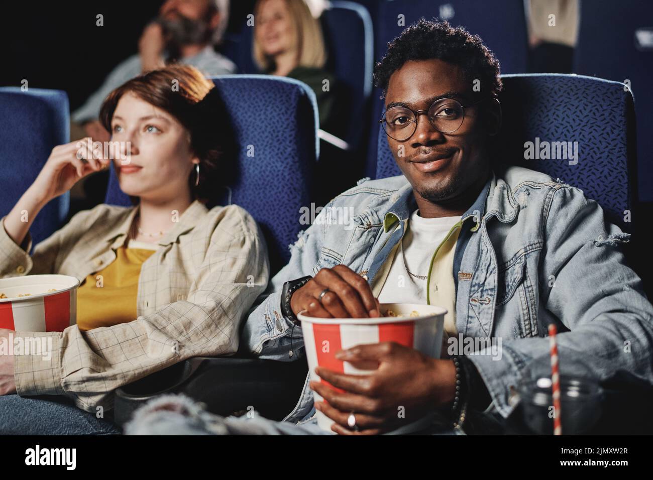 Captura selectiva de un alegre joven afroamericano con ropa informal sentado con su novia en el cine sonriendo a la cámara Foto de stock