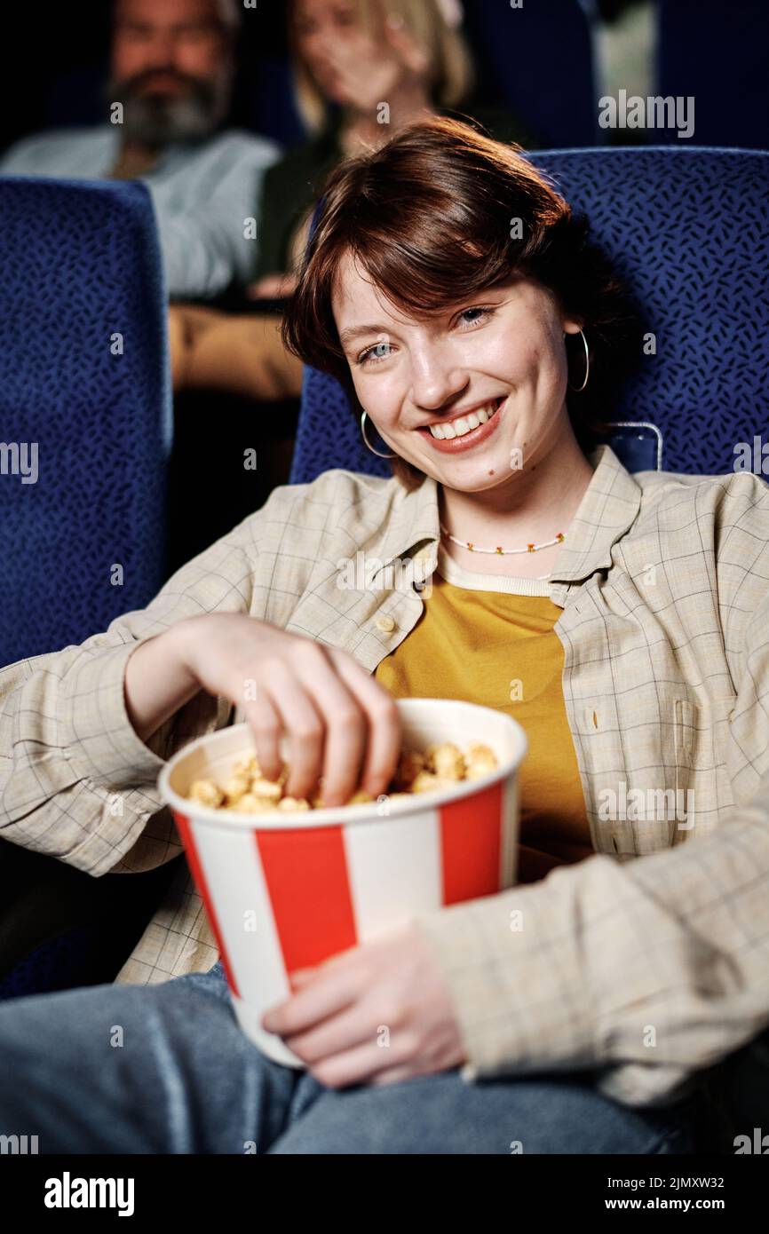 Retrato vertical medio de una encantadora joven con ropa informal sentada relajada comiendo palomitas de maíz y viendo una película mirando la cámara Foto de stock
