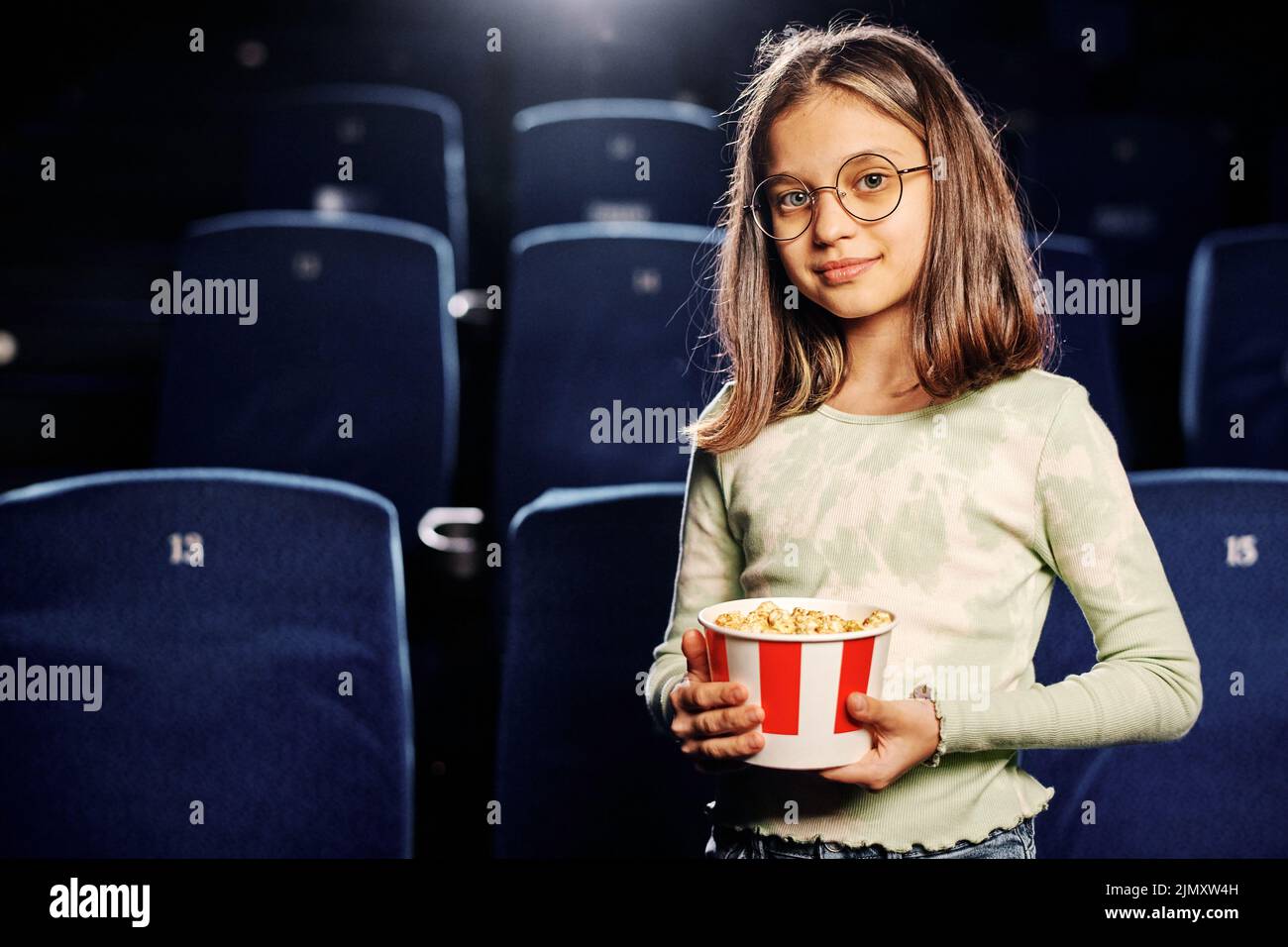 Retrato horizontal medio de una hermosa niña caucásica preadolescente sosteniendo un cubo de palomitas de maíz en el salón del cine contra los asientos azul marino Foto de stock