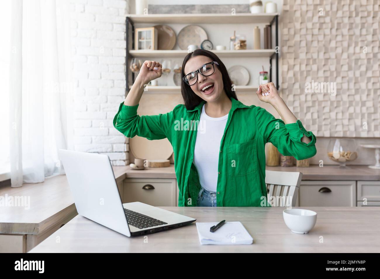 Retrato de una mujer de negocios feliz con gafas y chaqueta verde. Se sienta a la mesa en casa, trabaja con un portátil, está feliz, alzó las manos, recibió buenas noticias, sonríe. Foto de stock
