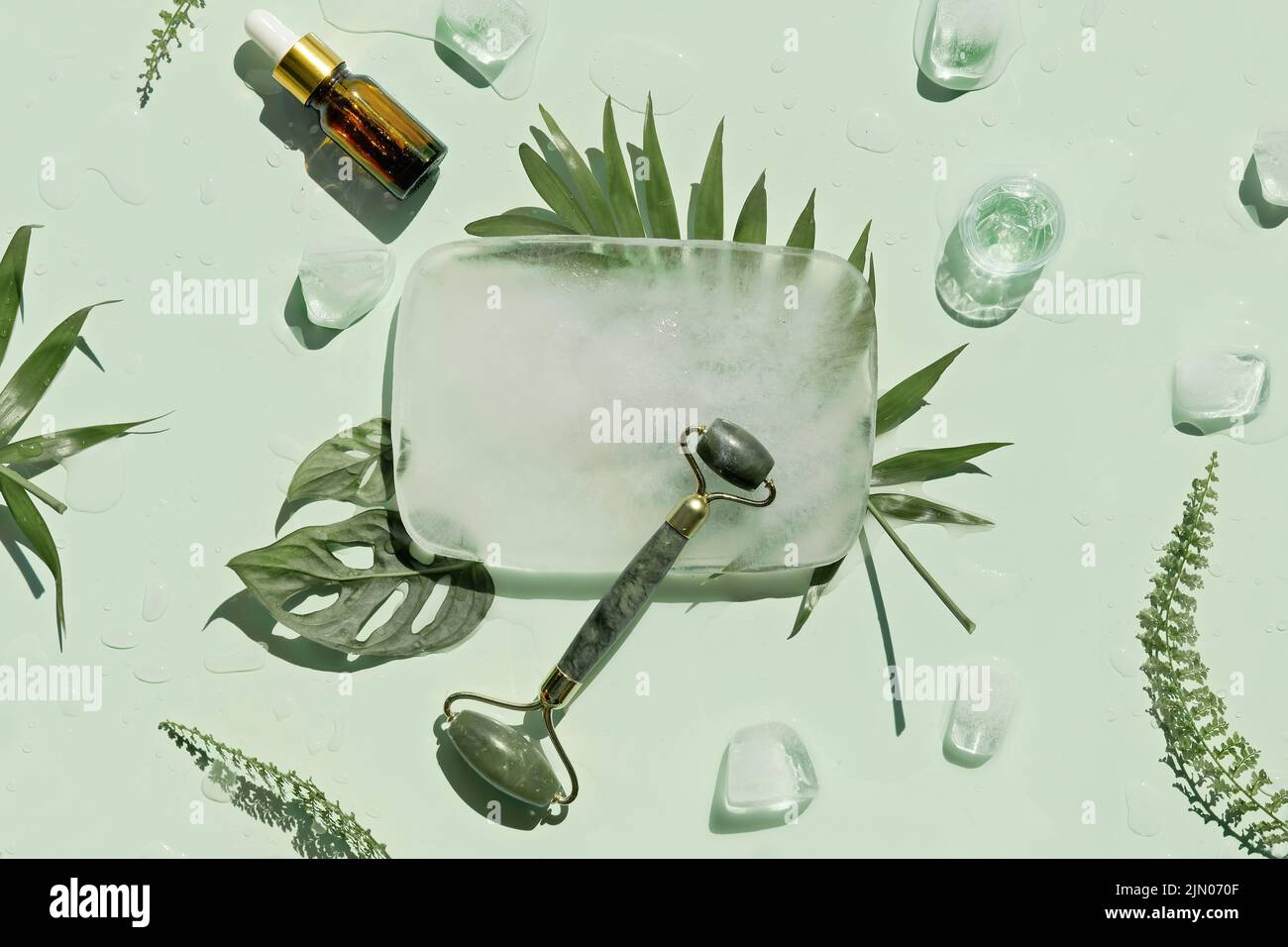 Rodillo facial de piedra de jade, aceite esencial y crema hecha a mano con cubitos de hielo y hojas exóticas de helecho de palma sobre fondo verde menta. Foto de stock
