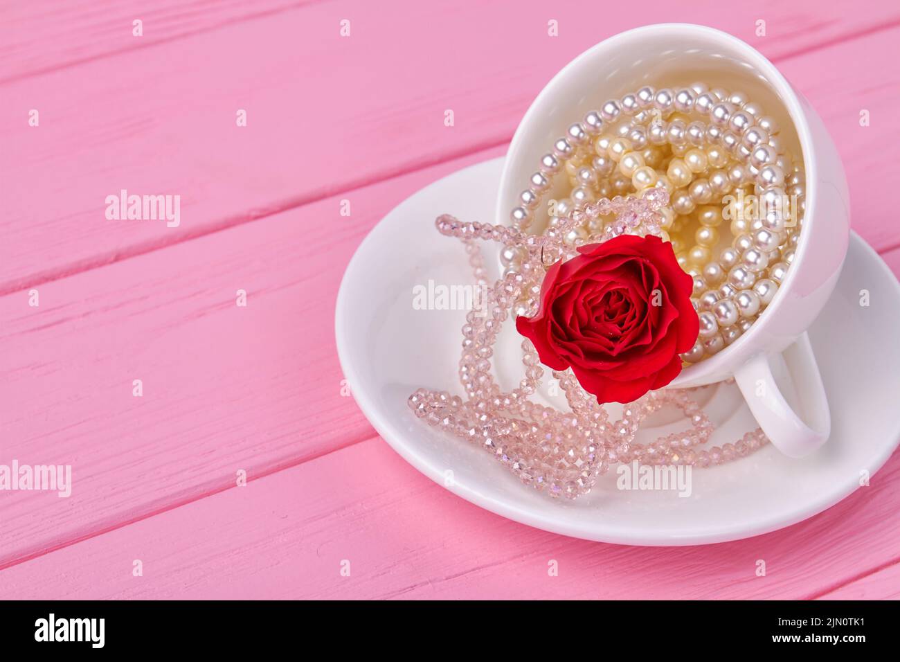 Copa blanca llena de collares de perlas y flores rojas. Escritorio de madera rosa. Foto de stock