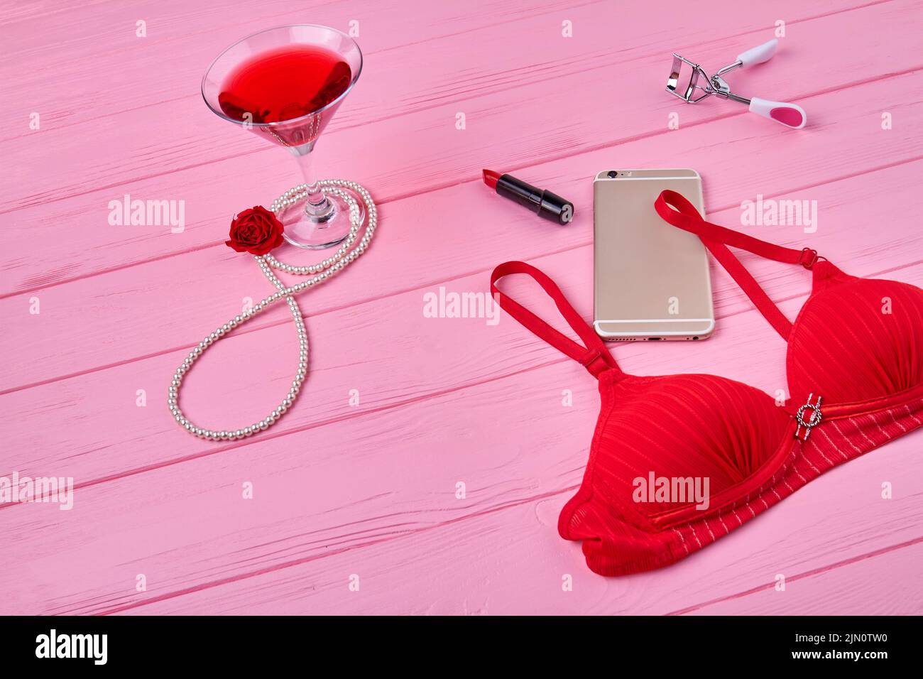 Sujetador rojo con smartphone y bebida alcohólica. Vista superior accesorios womans sobre escritorio de madera rosa. Foto de stock