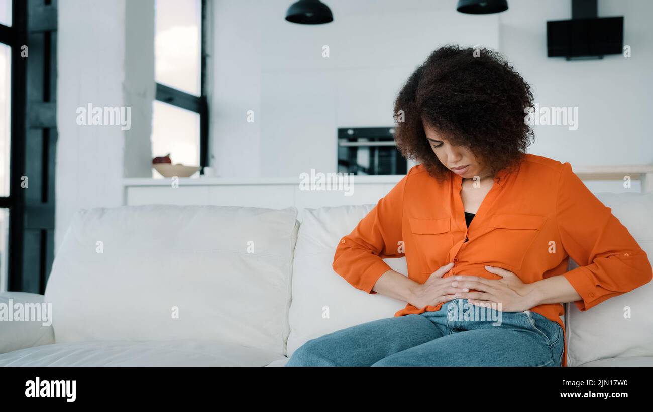 Mujer afroamericana estresada sentada en el sofá tocando vientre enferma niña biracial rizada que sufre de dolor de estómago. Señora infeliz sintiendo dolor Foto de stock