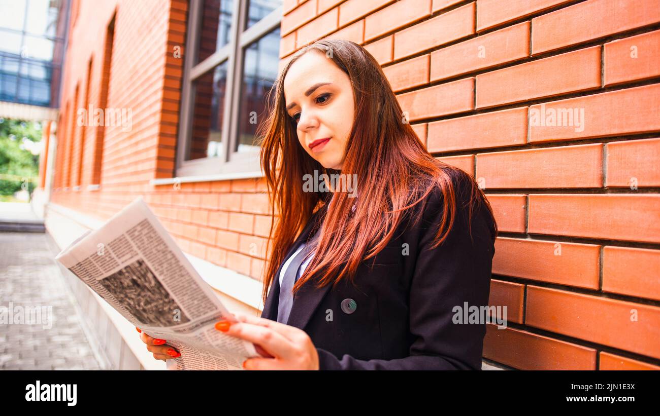 Mujer joven leyendo el periódico en la calle. Vista lateral de mujer joven con pelo largo en ropa informal de pie en la calle y leyendo el periódico. Foto de stock