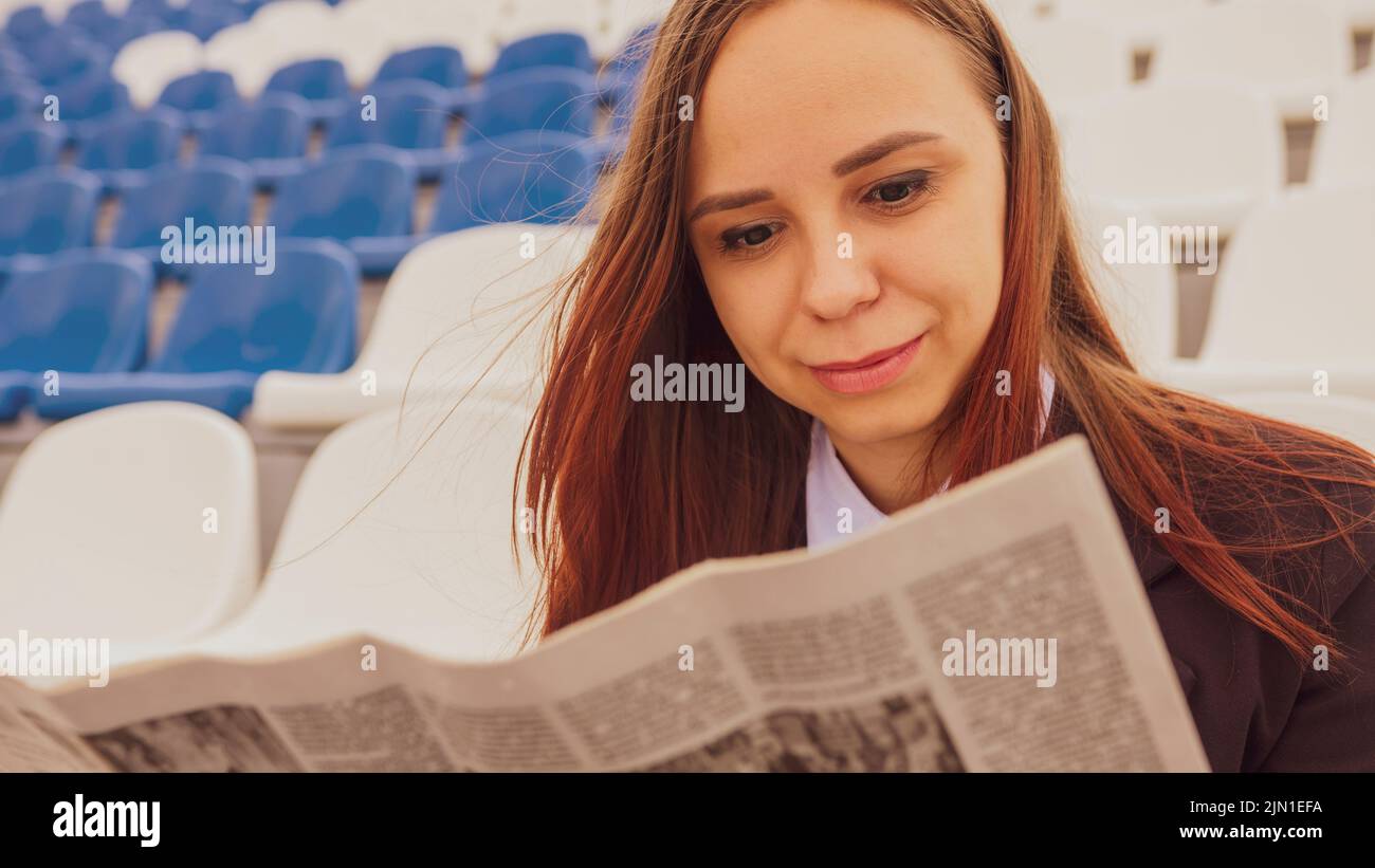 Una mujer en un traje de negocios lee un periódico mientras está sentada en el estadio. Foto de stock