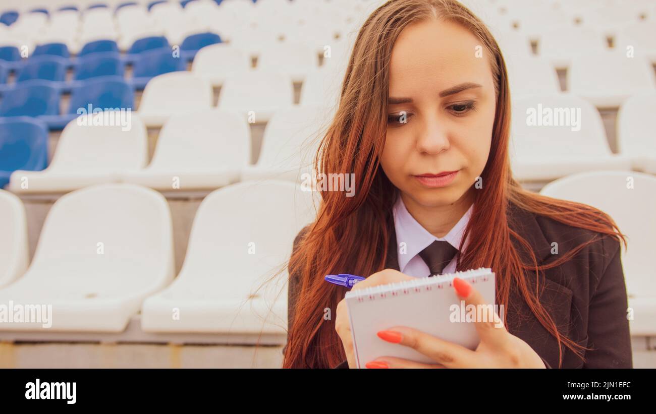 Mujer joven con bloc de notas, pluma sentada en gradas del estadio solo. Una periodista escribía notas durante el entrenamiento deportivo en el estadio de la calle Foto de stock