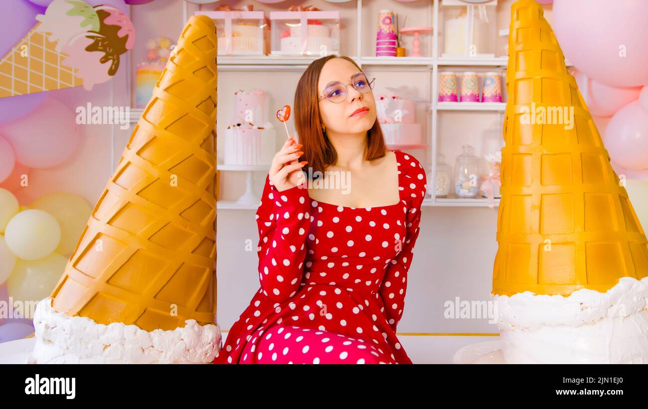 Mujer comiendo pirup en confitería. La hembra contenta mira lejos mientras que come el lollipop dulce en la tabla con los conos decorativos grandes del helado en luz Foto de stock