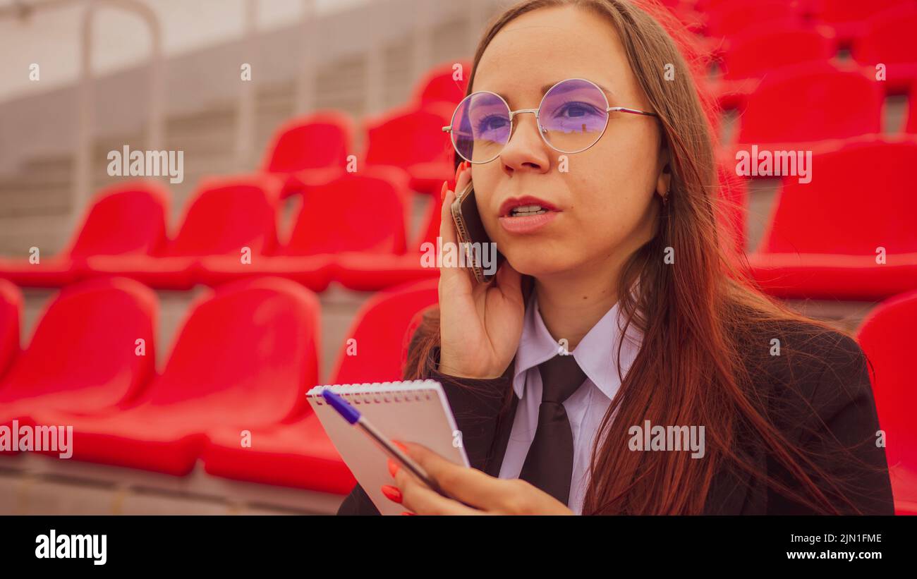 Mujer joven en vasos con bloc de notas, bolígrafo hablando en el teléfono móvil, sentado en gradas estadio Foto de stock