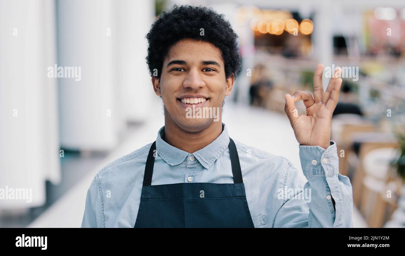 Joven hombre africano americano trabajador camarero vendedor en delantal hombre pequeño negocio dueño de la cafetería restaurante tienda mirando la cámara con una sonrisa amistosa Foto de stock