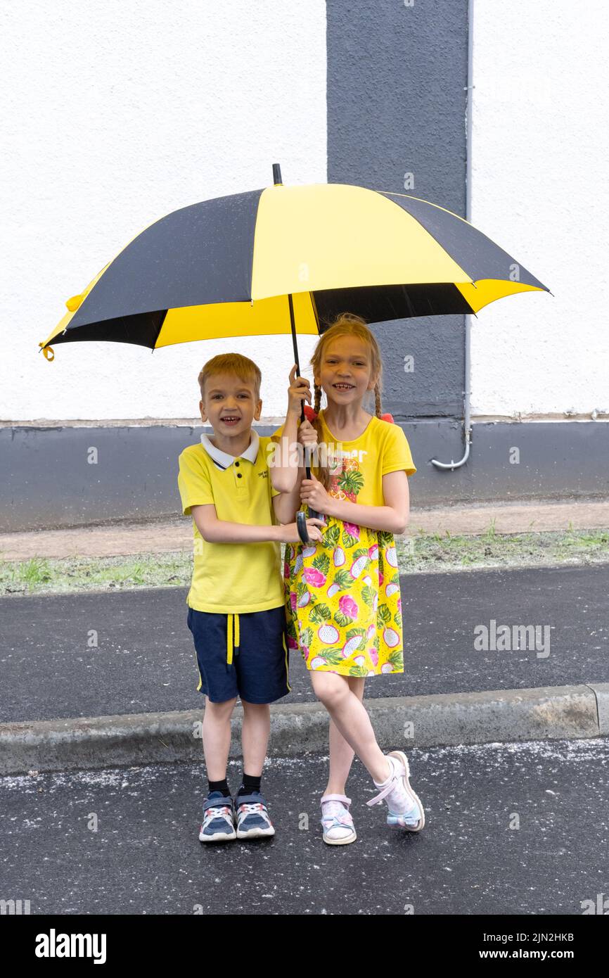 Retrato de dos niños sonrientes, un niño y una niña, de pie bajo una sombrilla en el parque en verano. Los niños se regocijan en la lluvia estival. Foto de stock