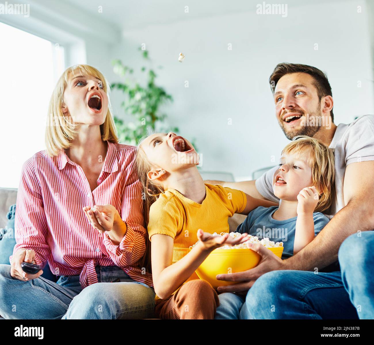 Familia divirtiéndose viendo la televisión y comiendo jugando con palomitas en el sofá de la casa Foto de stock