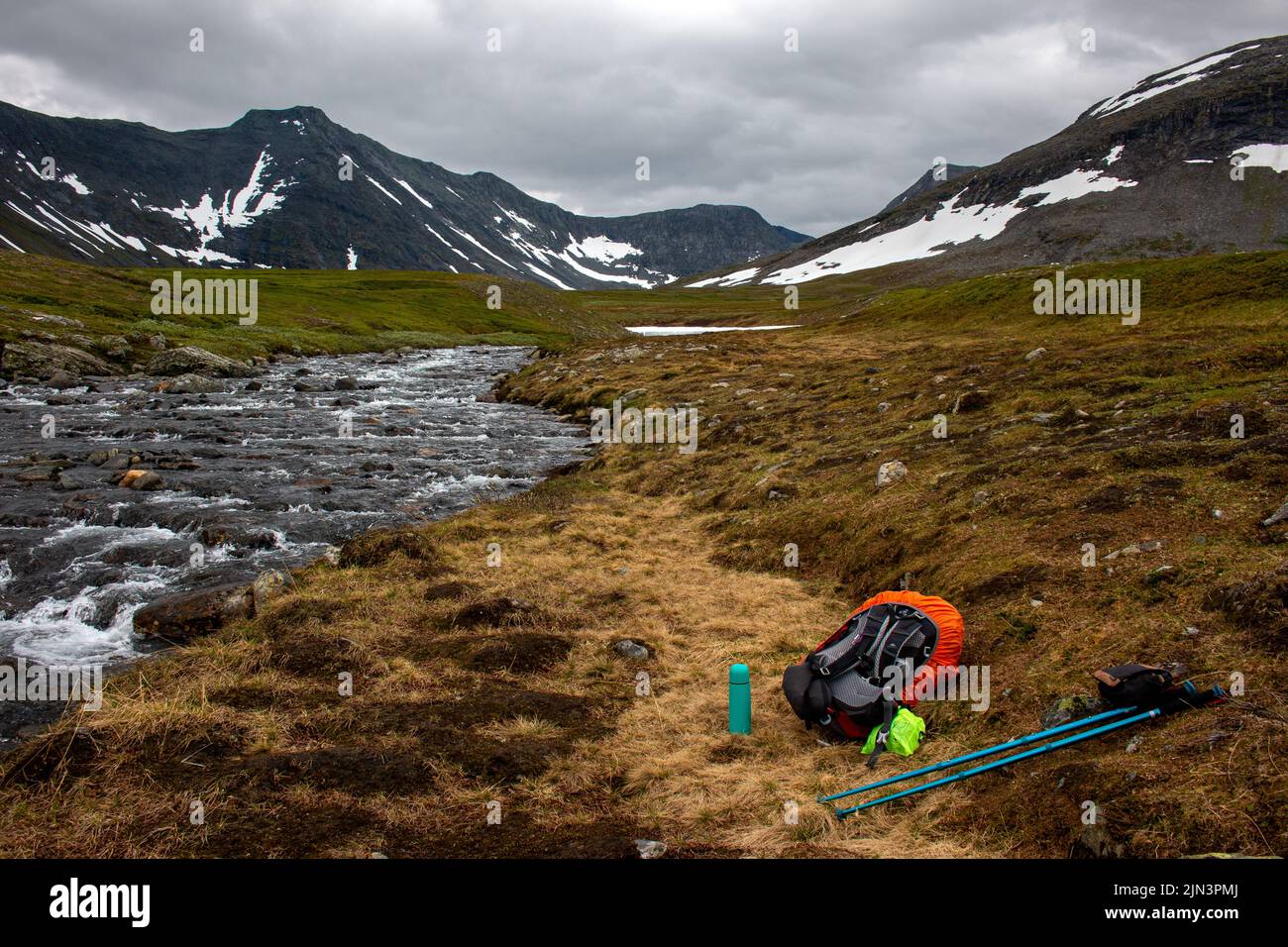 Una mochila, bastones de senderismo, y un termo junto a un arroyo que viene de la nieve que se derrite en la cima de las montañas Sylarna, Noruega Foto de stock