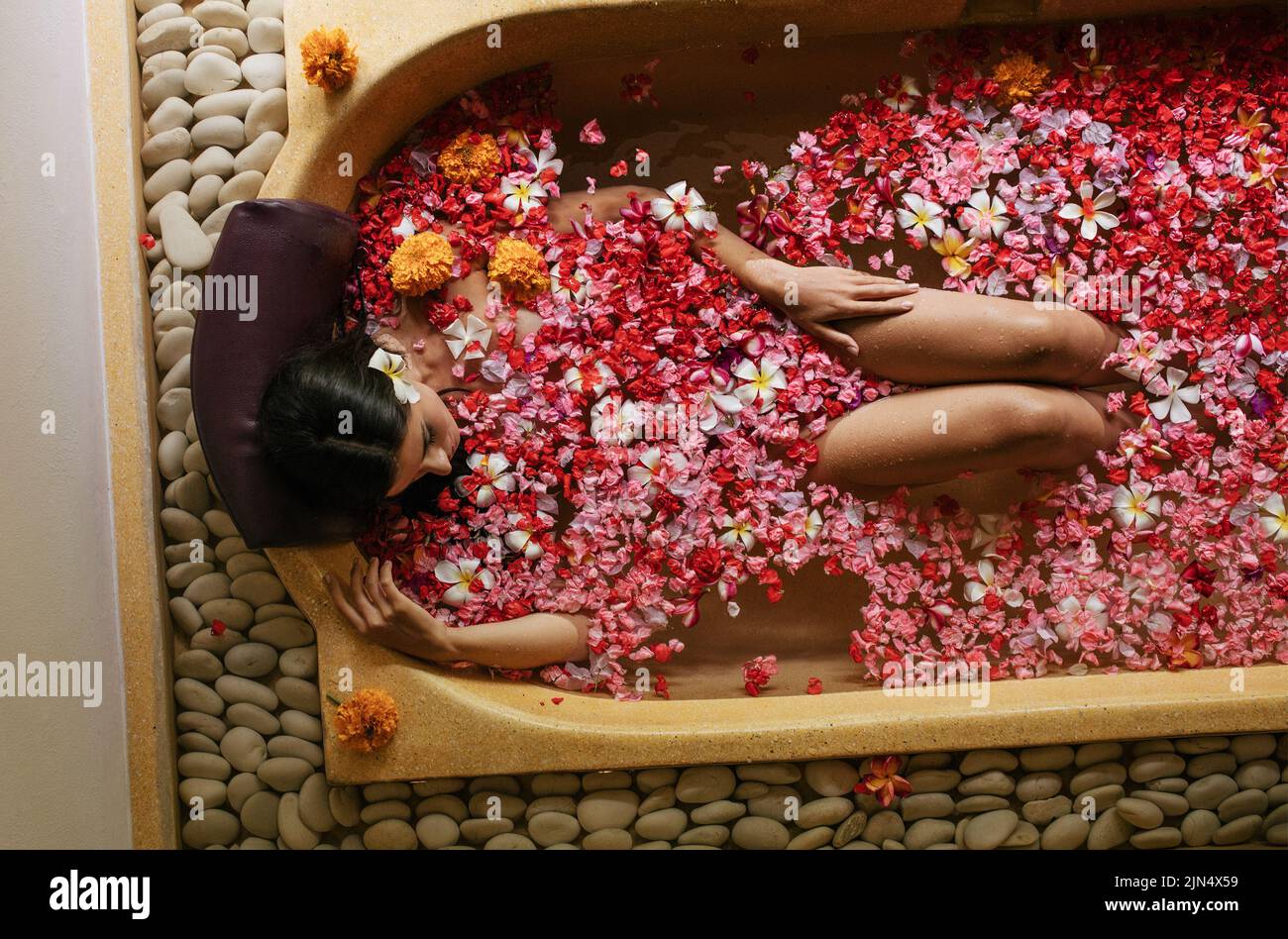 Vista superior de mujer tomando baño con flores flotando en el agua. Mujer en la bañera con pétalos en el spa. Foto de stock