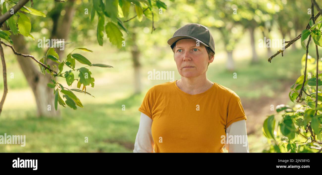 Retrato de una trabajadora campesina con camiseta naranja y sombrero de camionero en un huerto de nogal, enfoque selectivo Foto de stock