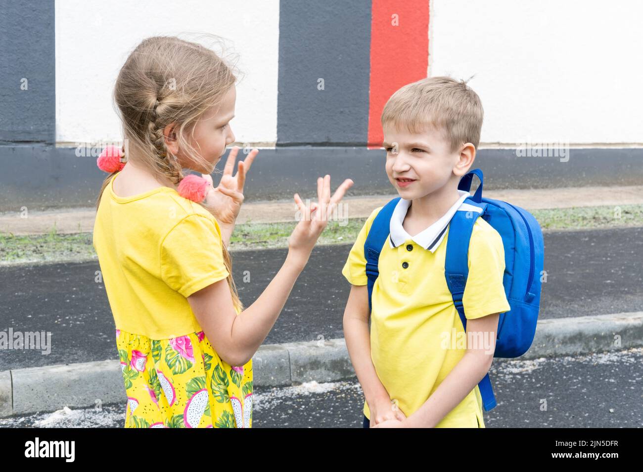 De vuelta a la escuela. La hermana mayor le dice gestos al hermano menor con una mochila escolar. Los niños se comunican antes de la escuela. Foto de stock