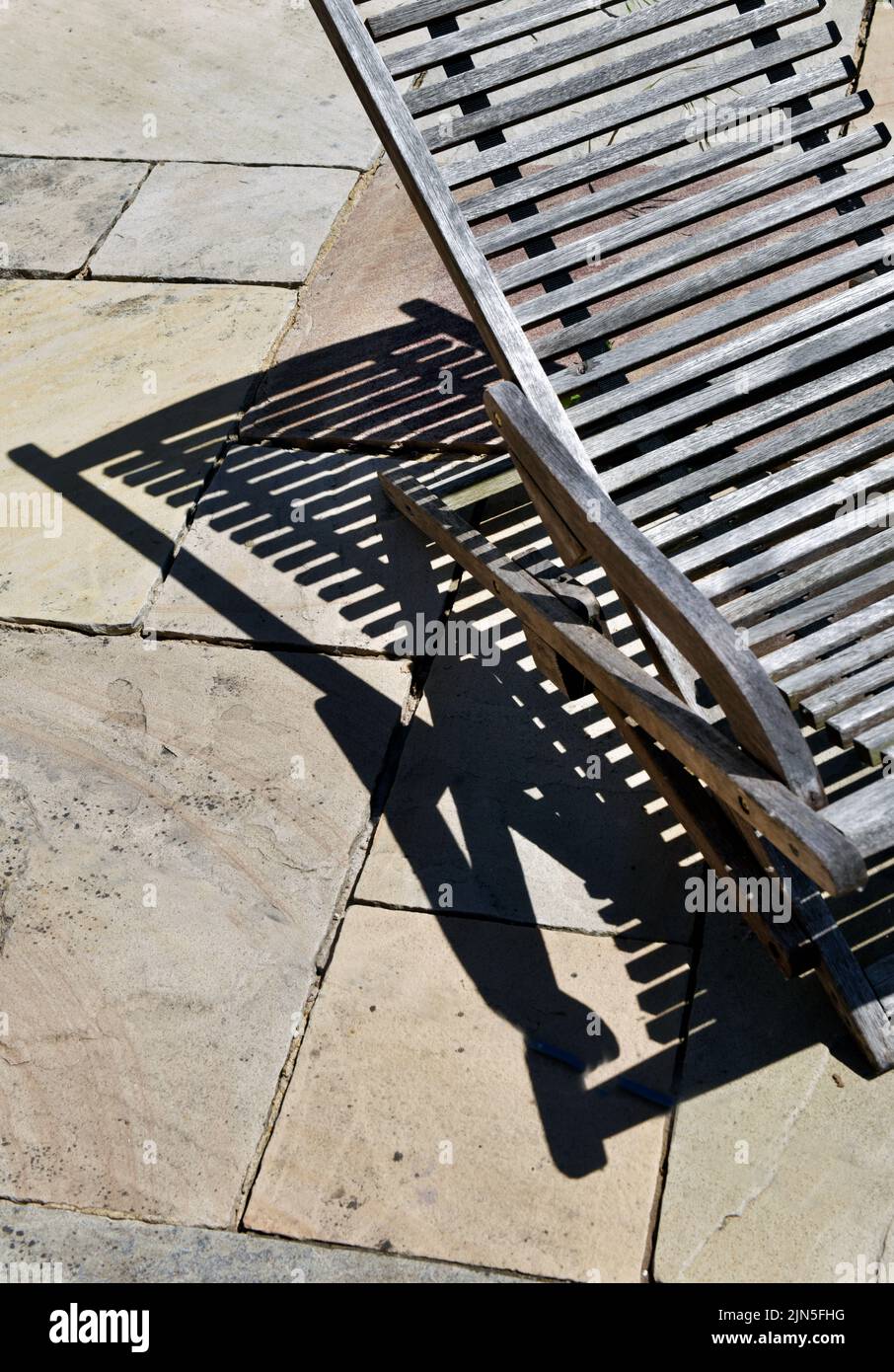 una tumbona de madera bien desgastada que proyecta una fuerte sombra sobre las losas de pavimentación Foto de stock