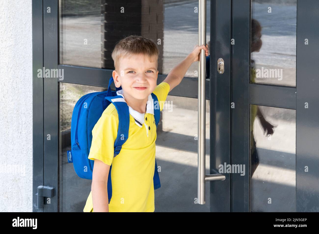 El muchacho caucásico con confianza sale de casa para su primer día en el preescolar. Un lindo niño con una mochila de la escuela está de pie cerca de la puerta principal de la Foto de stock
