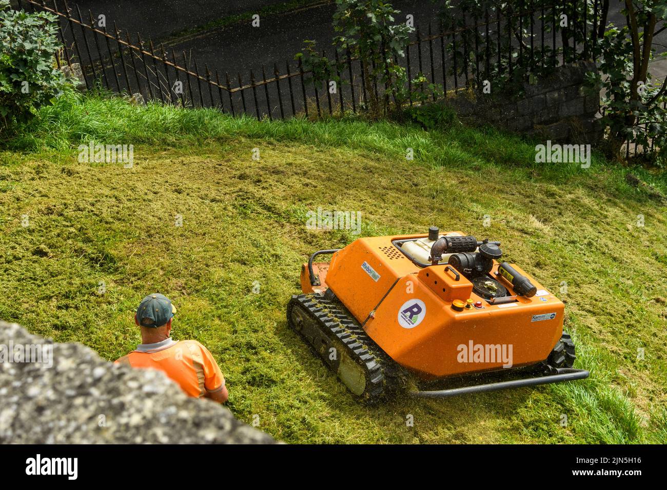Hierba pendiente segada por cortacésped robótico naranja controlado a distancia (KommTek RoboFlail) y trabajador en hi-vis - York Walls, Yorkshire, Inglaterra, Reino Unido. Foto de stock