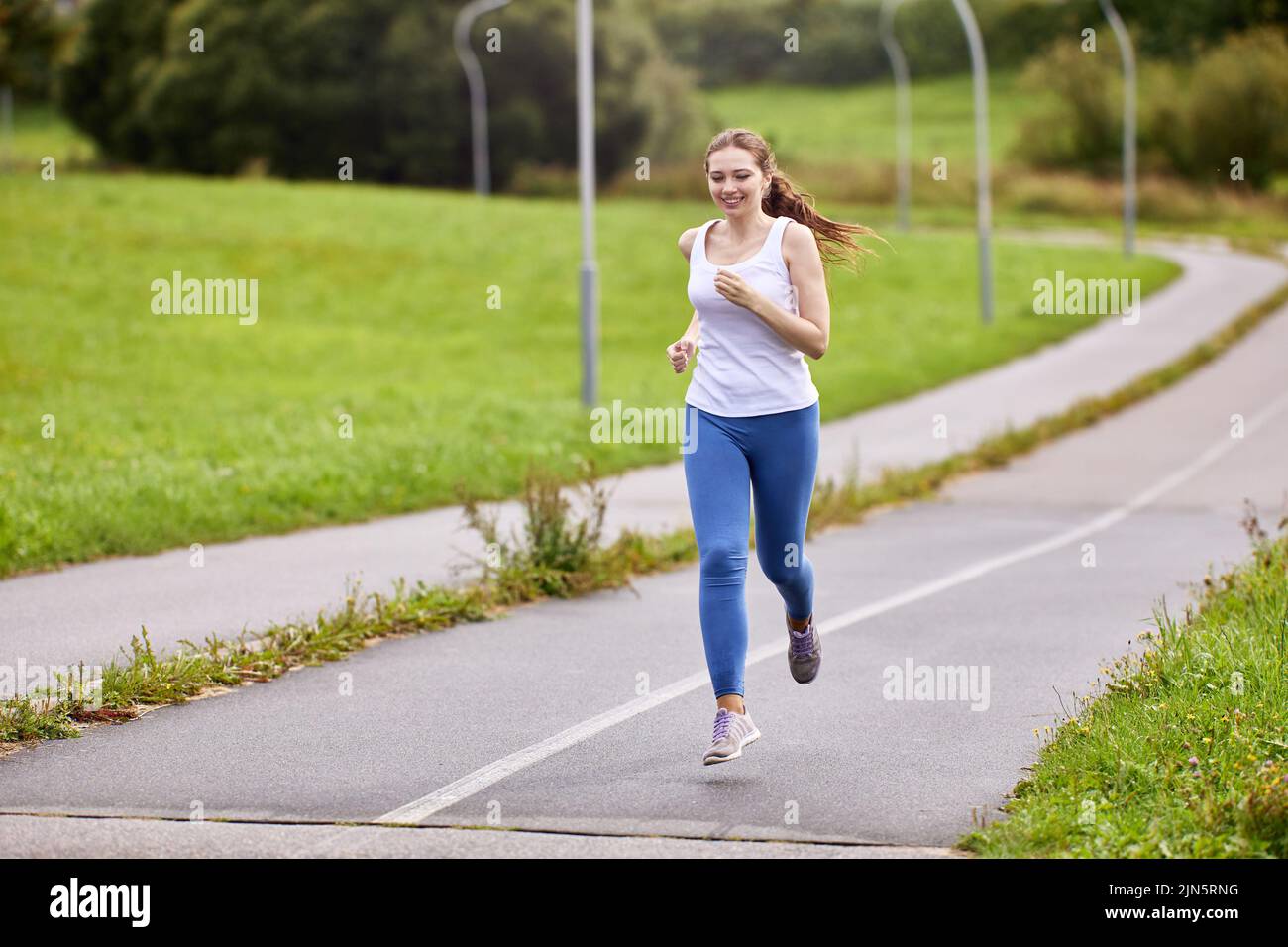 Una corredora con pantalón y leggings blancos hace correr por la mañana por la pista de ciclismo en el parque de la ciudad durante el verano. Foto de stock