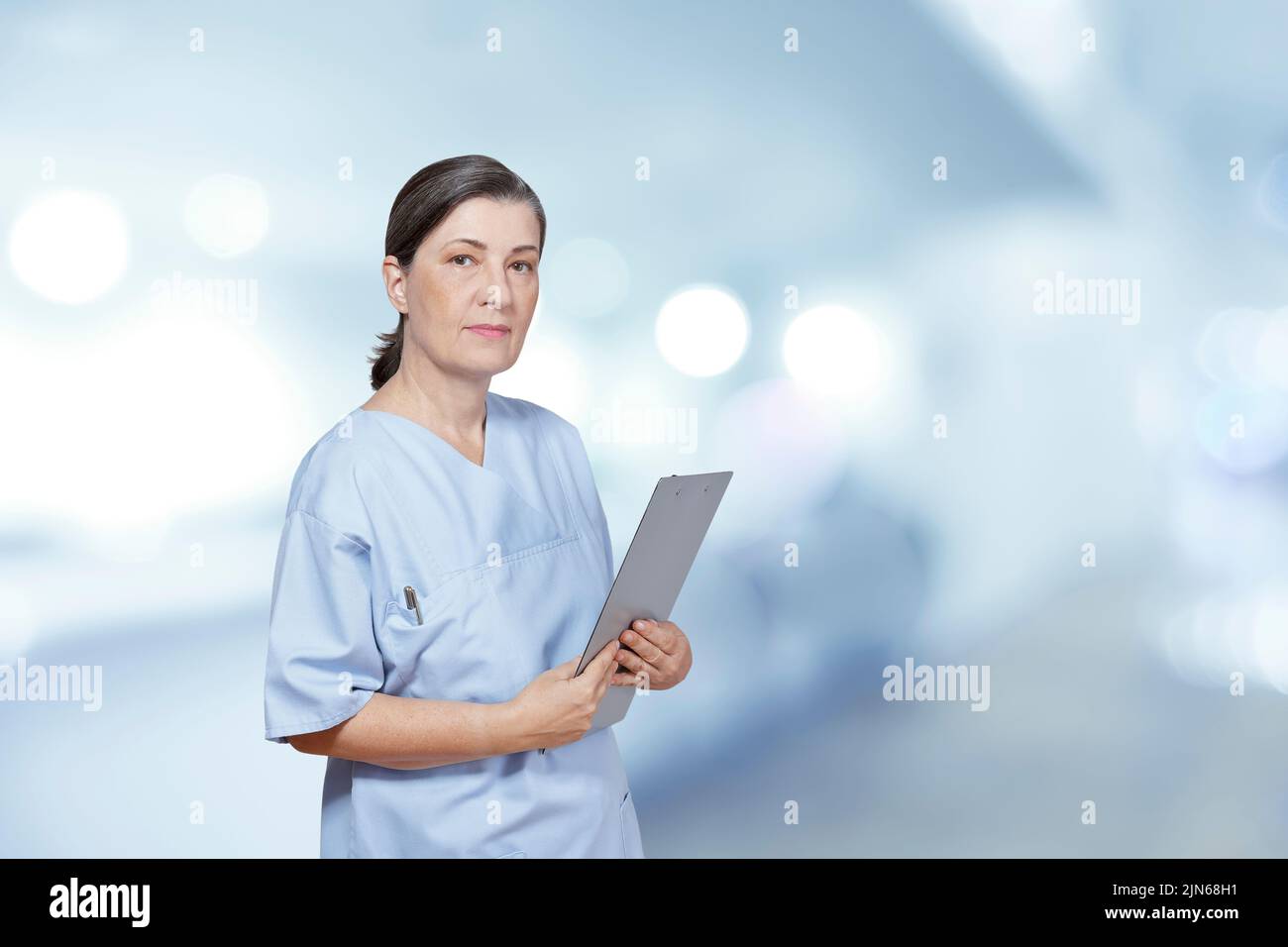 Retrato de una enfermera más vieja en un matorral azul que sostiene un sujetapapeles, fondo interior del hospital, efecto de filtro azul. Foto de stock