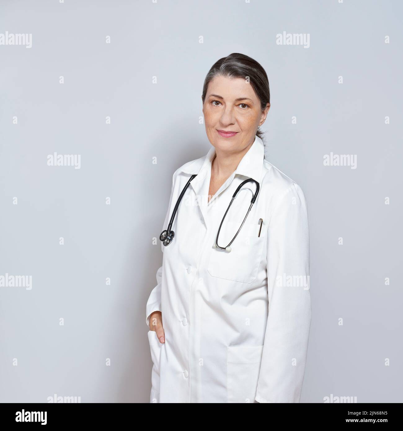 Doctora de mediana edad con bata de laboratorio blanca, sonriente, sobre fondo gris o gris neutro, copia o espacio de texto. Foto de stock