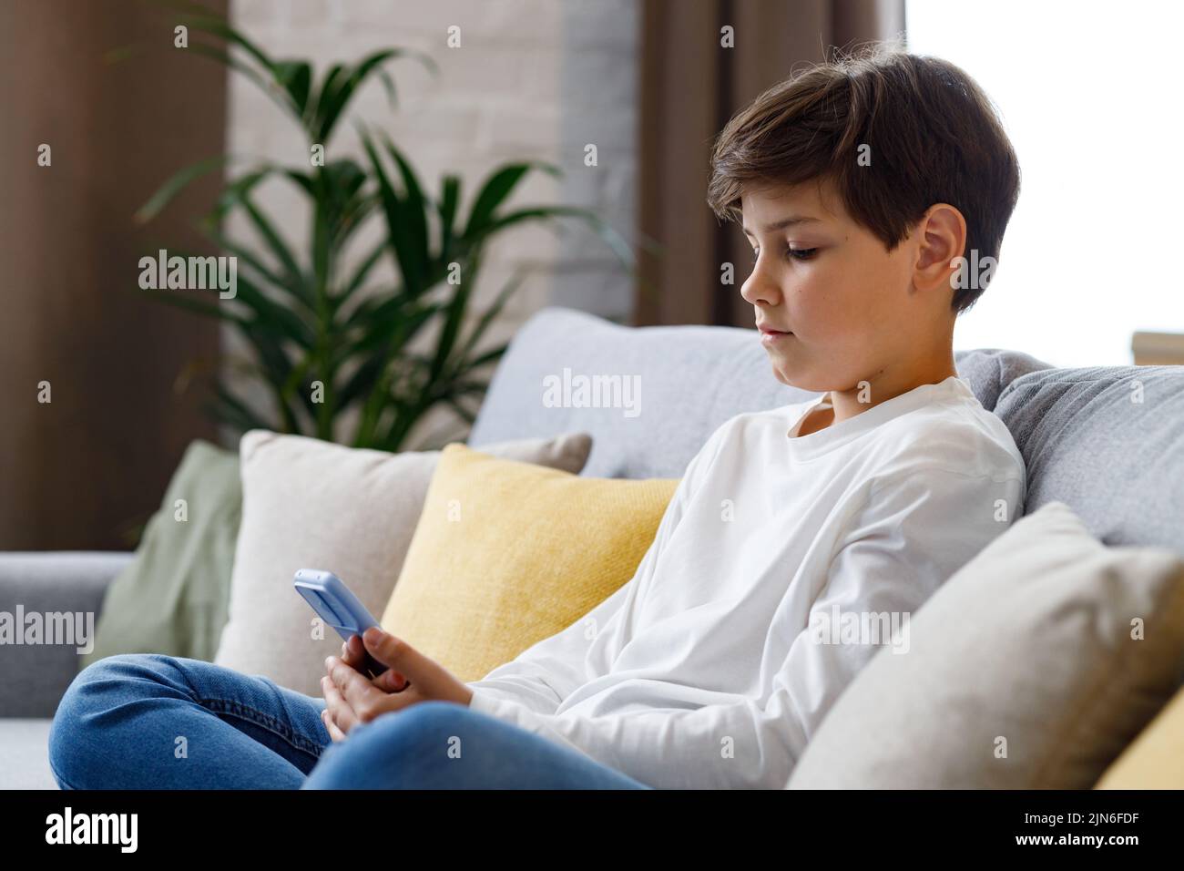 Retrato de joven lindo niño sosteniendo smartphone sentado en el sofá. Adolescente en casa eligiendo su música favorita, enviando mensajes de texto, navegando por Internet, viendo Foto de stock