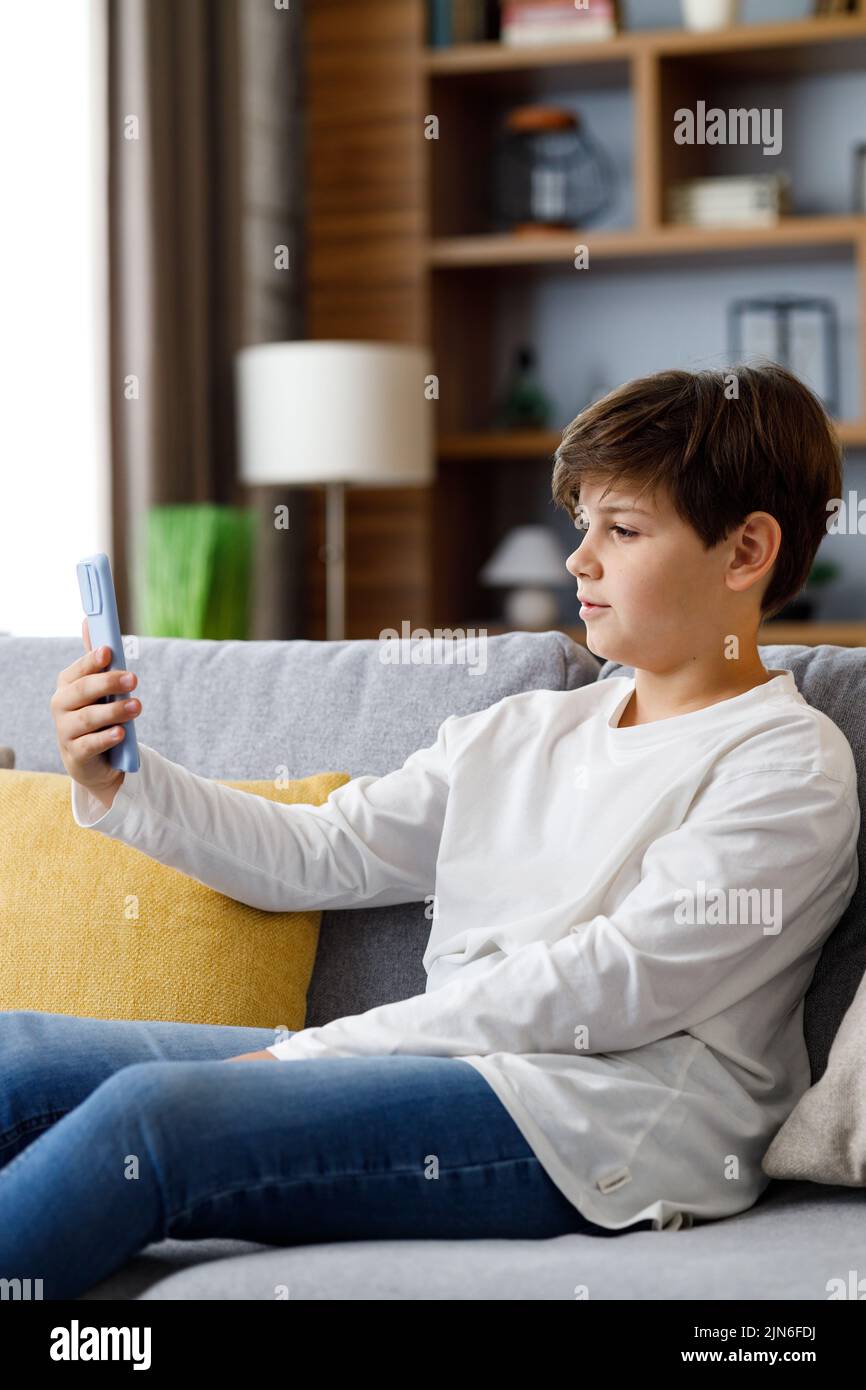 Retrato de joven lindo niño sosteniendo smartphone sentado en el sofá. Adolescente en casa eligiendo su música favorita, enviando mensajes de texto, navegando por Internet, viendo Foto de stock