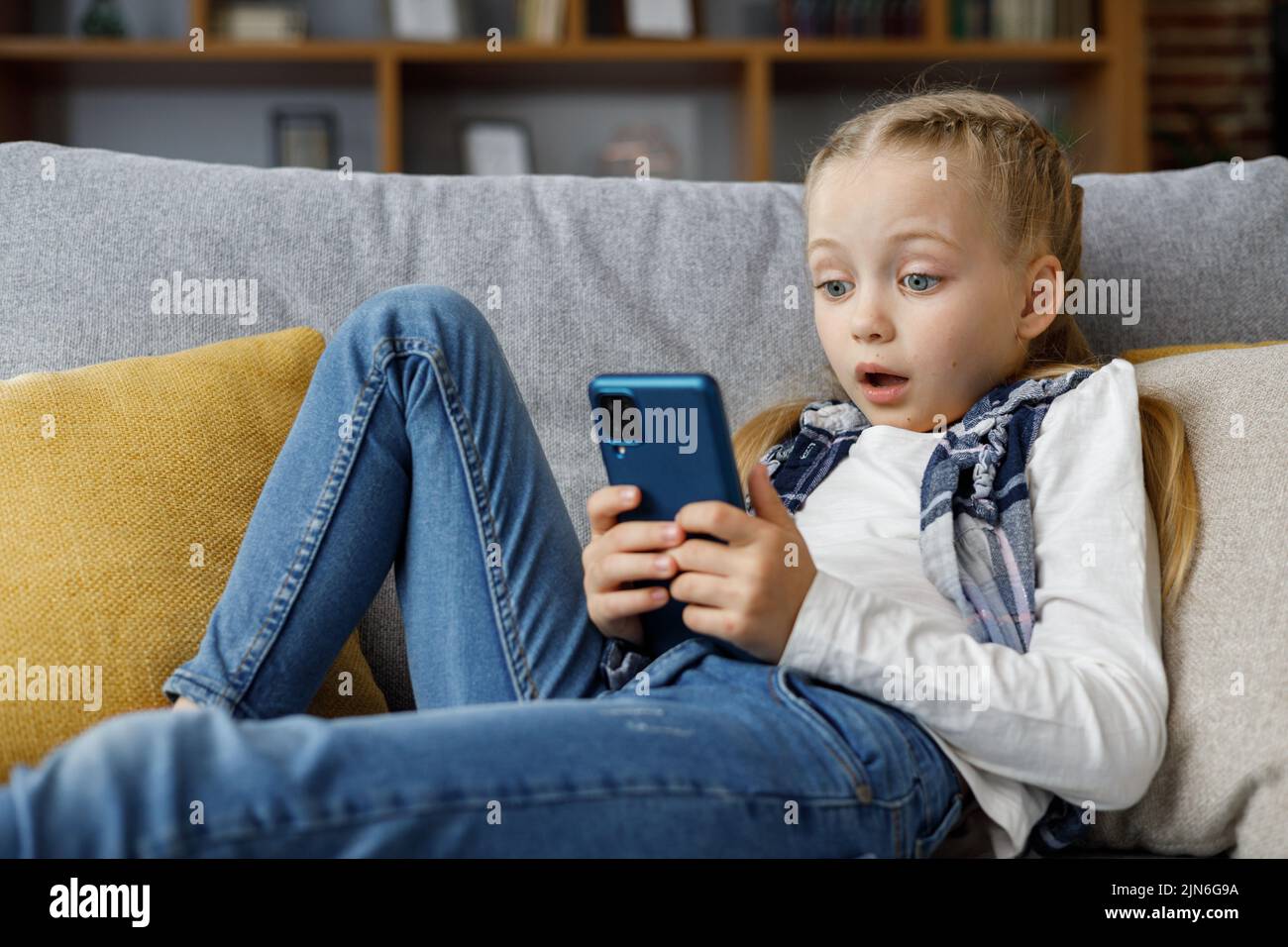 Sorprendida pequeña chica linda sosteniendo smartphone y descansando en el sofá en casa. Pasar tiempo en las redes sociales, mensajes de texto, navegar por Internet Foto de stock