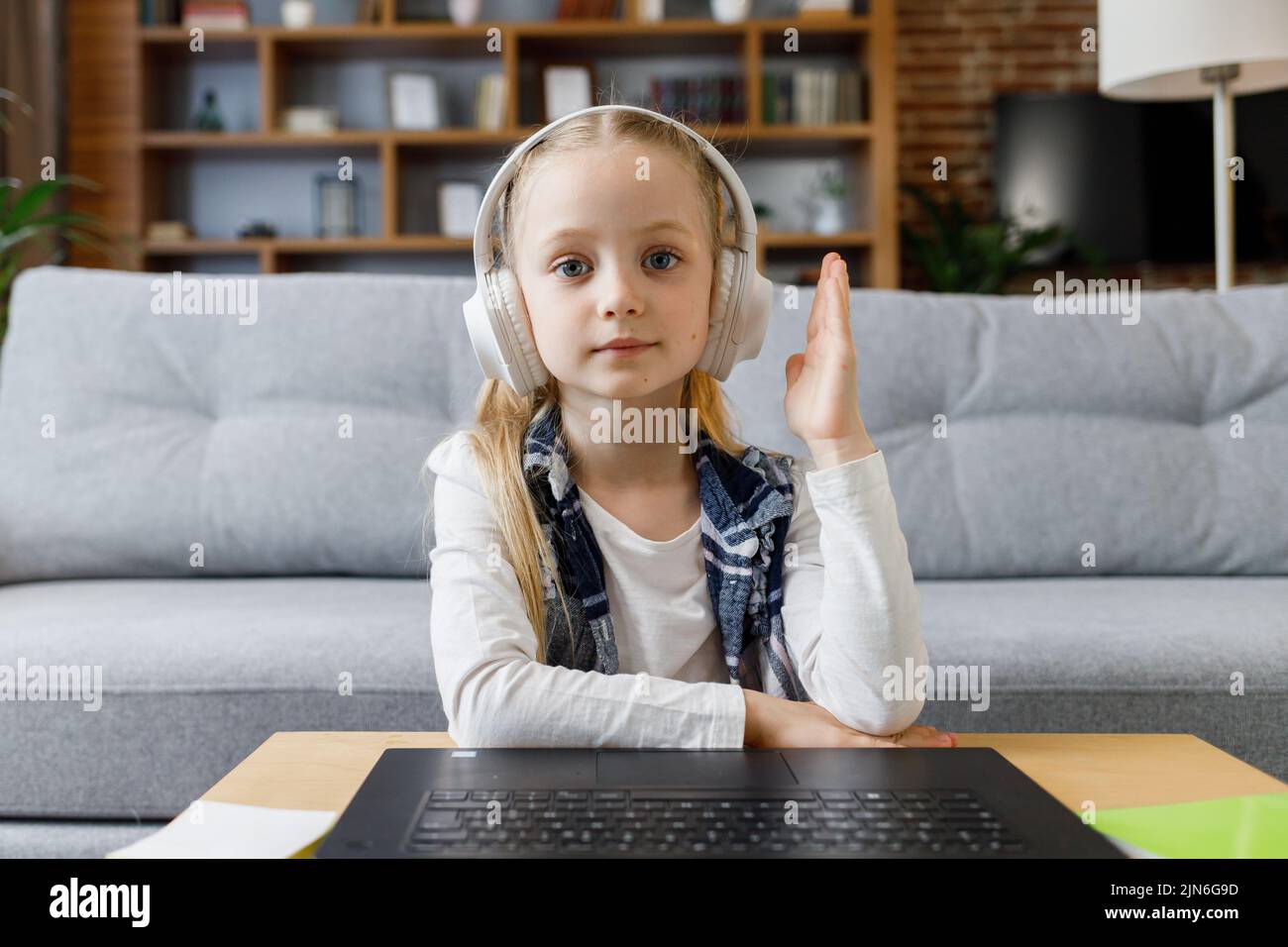 Primaria escolar con auriculares tiene lección en línea, video llamada con el profesor, levantar la mano para responder a la pregunta. Lindo niño estudiando en casa Foto de stock