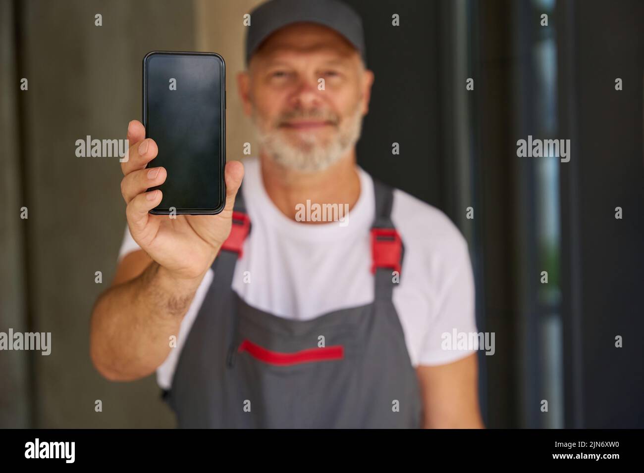 Un constructor sonriente con gorro y monos muestra el teléfono móvil Foto de stock