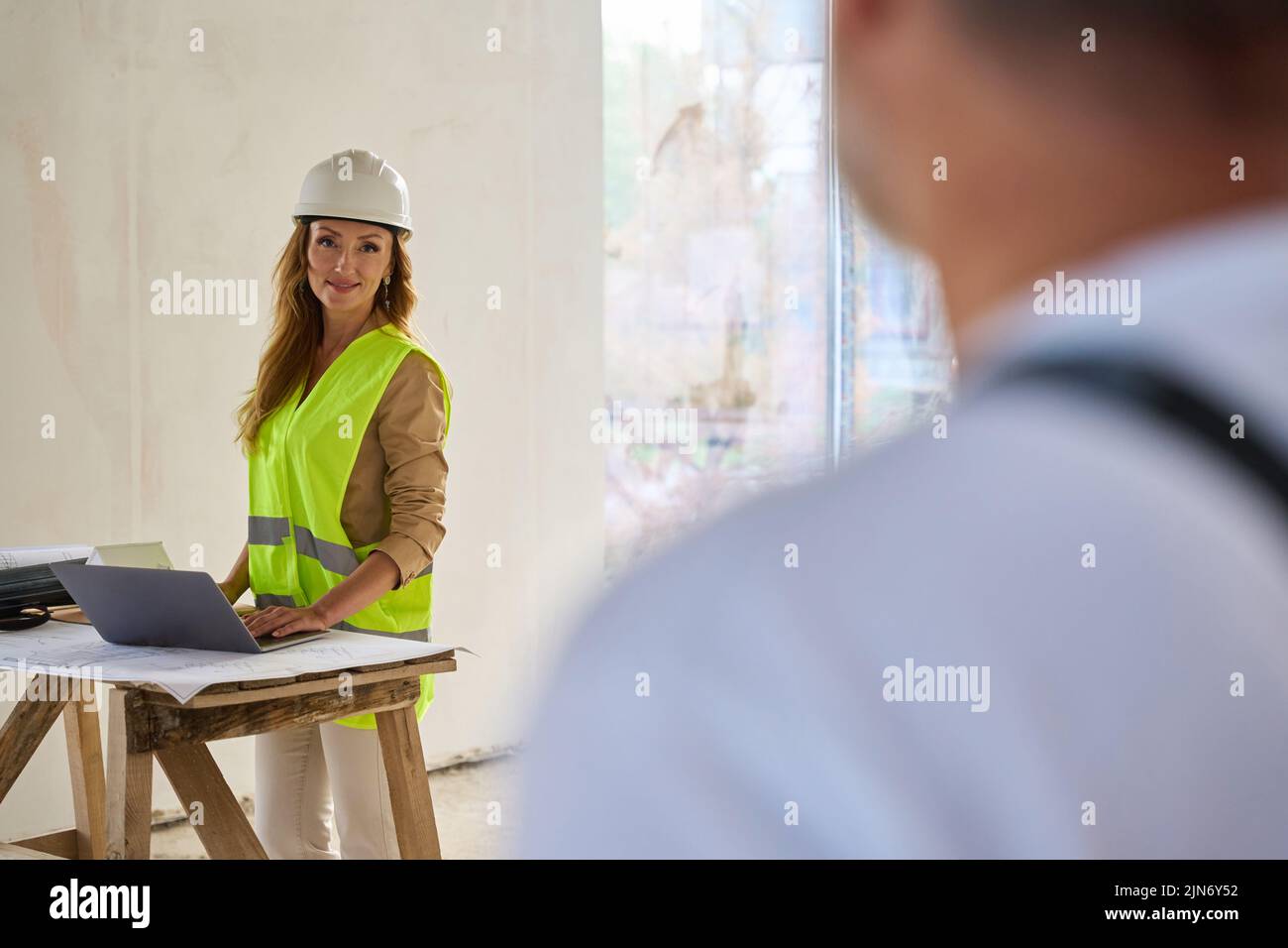 Constructor mirando al agente inmobiliario parado cerca de la mesa con el ordenador portátil Foto de stock