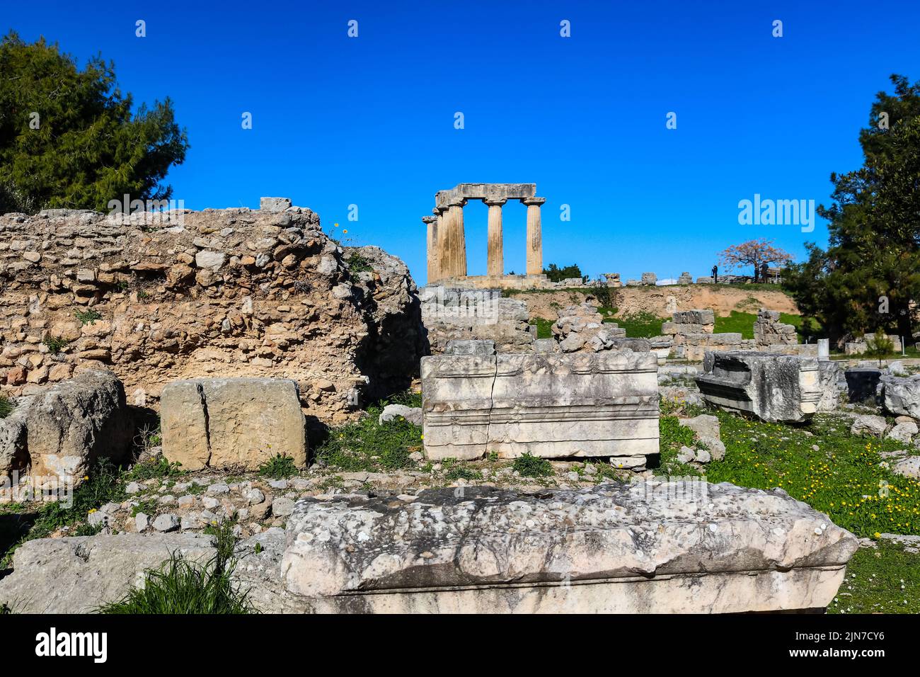 Templo de Apolo en la antigua Corinto Grecia visto desde abajo del hll en las ruinas excavadas con turistas inidentificables tomando fotos arriba y y. Foto de stock
