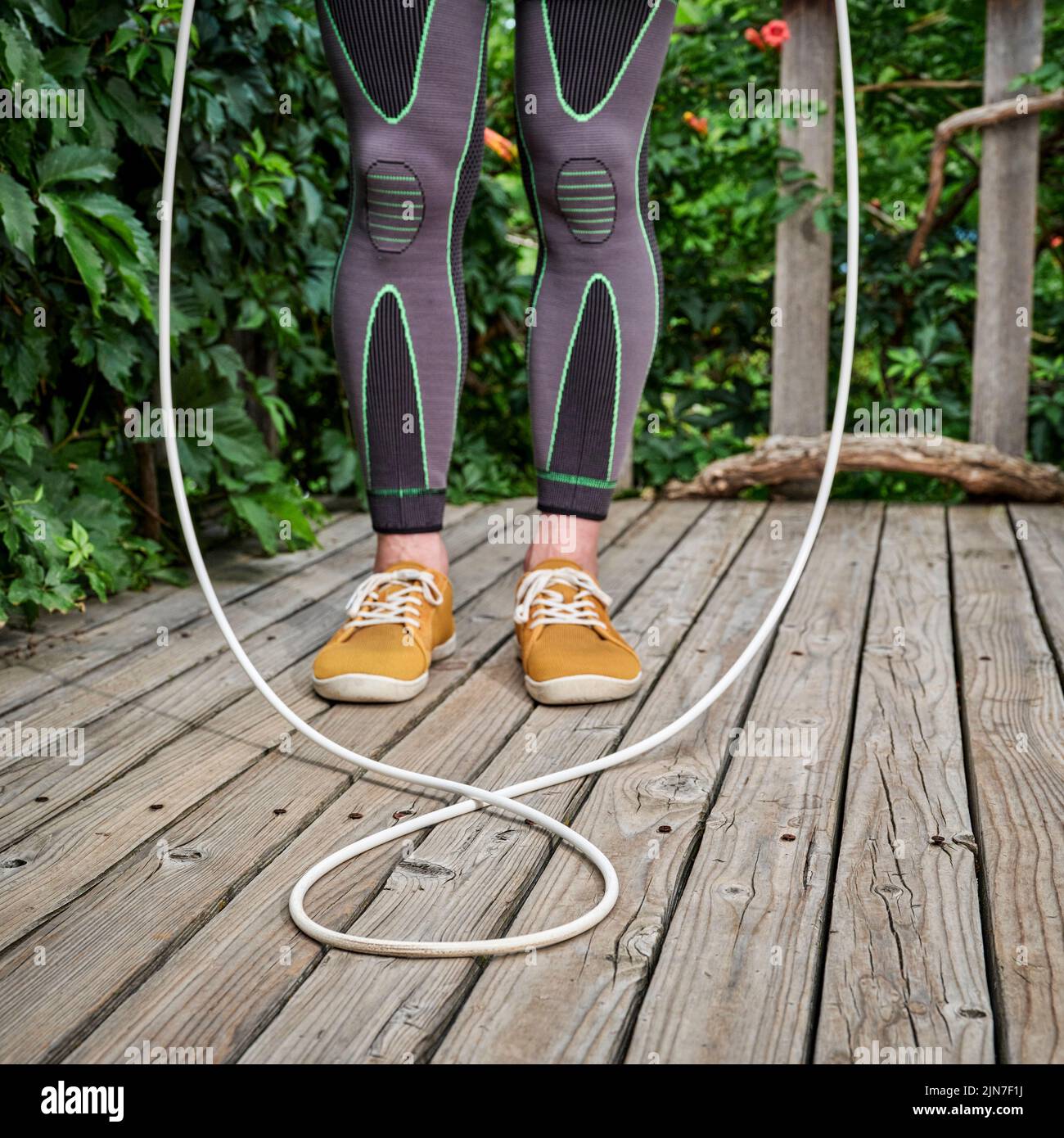 entrenamiento con una cuerda de salto pesada - piernas masculinas en medias de compresión en una cubierta de madera, concepto de fitness patio trasero Foto de stock