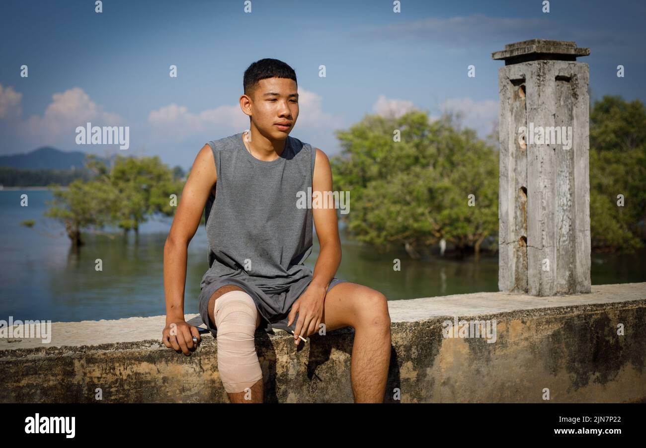 Un adolescente tailandés con una camiseta gris y una pierna vendada, se sienta sobre un dique con un cigarrillo en la mano con el mar azul y árboles verdes detrás Foto de stock